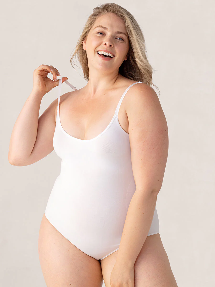 Blonde woman exuding joy in a 3XL white essentials scoop bodysuit, cheerfully holding up the right shoulder strap.